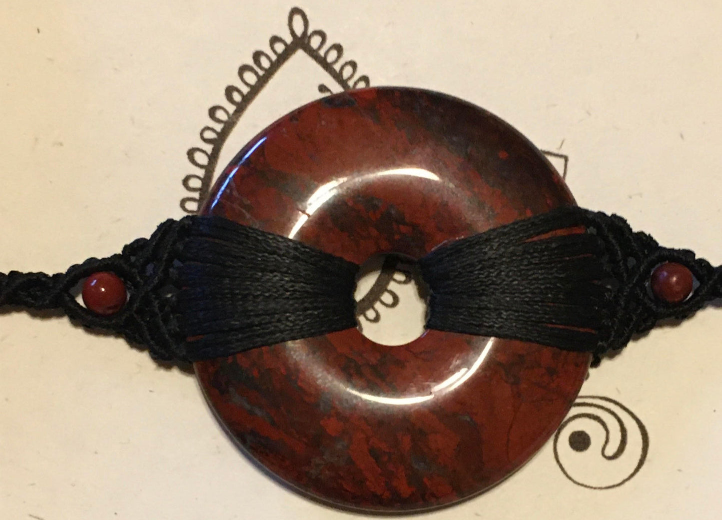 Red Jasper Hand Knotted Macrame Bracelet Artist: Erin Abraham Hand knotted macrame bracelet with Red Jasper gemstone Red Jasper donut occupies the center of this unique necklace 2" across stone Made with high quality black nylon cord There are no glues or other adhesives used in the construction of this necklace The bracelet features an adjustable closure, allowing it to be worn around various size wrists Each piece is a one-of-a-kind and will never be duplicated