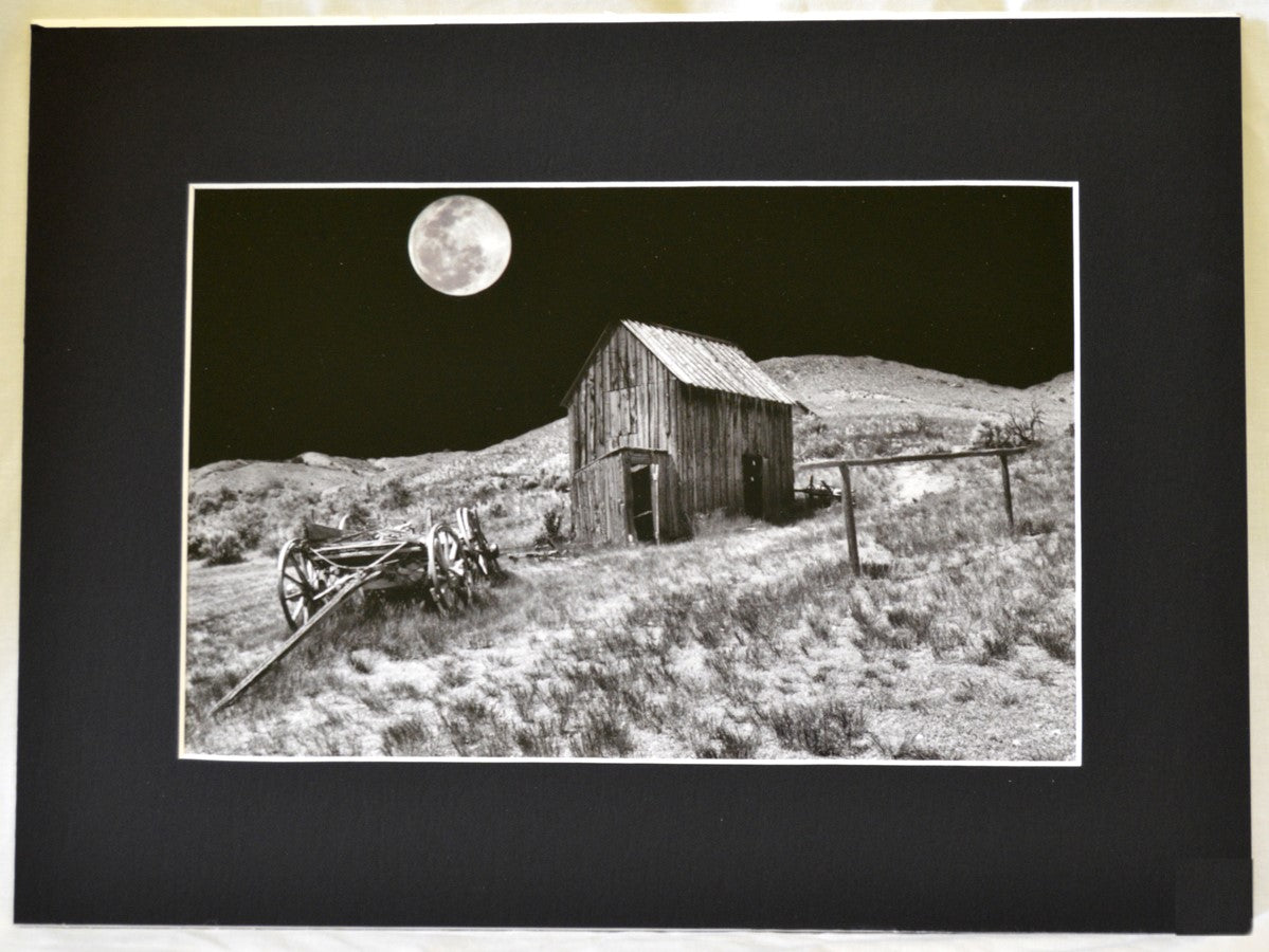 Matted Black and White photograph of an old barn, old wagon, and a full moon The image was taken in Bannack State Park, Montana 2" wide black matting with white edging around the print 12" long x 8" high print 16" long x 12" high as matted In a plastic sleeve and on sturdy foam board for added protection Ready for a frame or to be displayed on an easel
