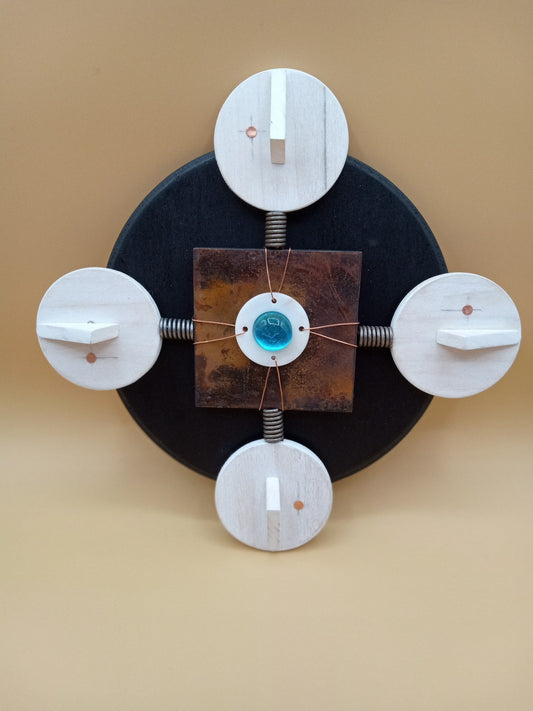 Round black board with square metal plaque with blue glass bead in center  On each side of plaque is a head with one copper eye  The shape and faces represent humanity uniting