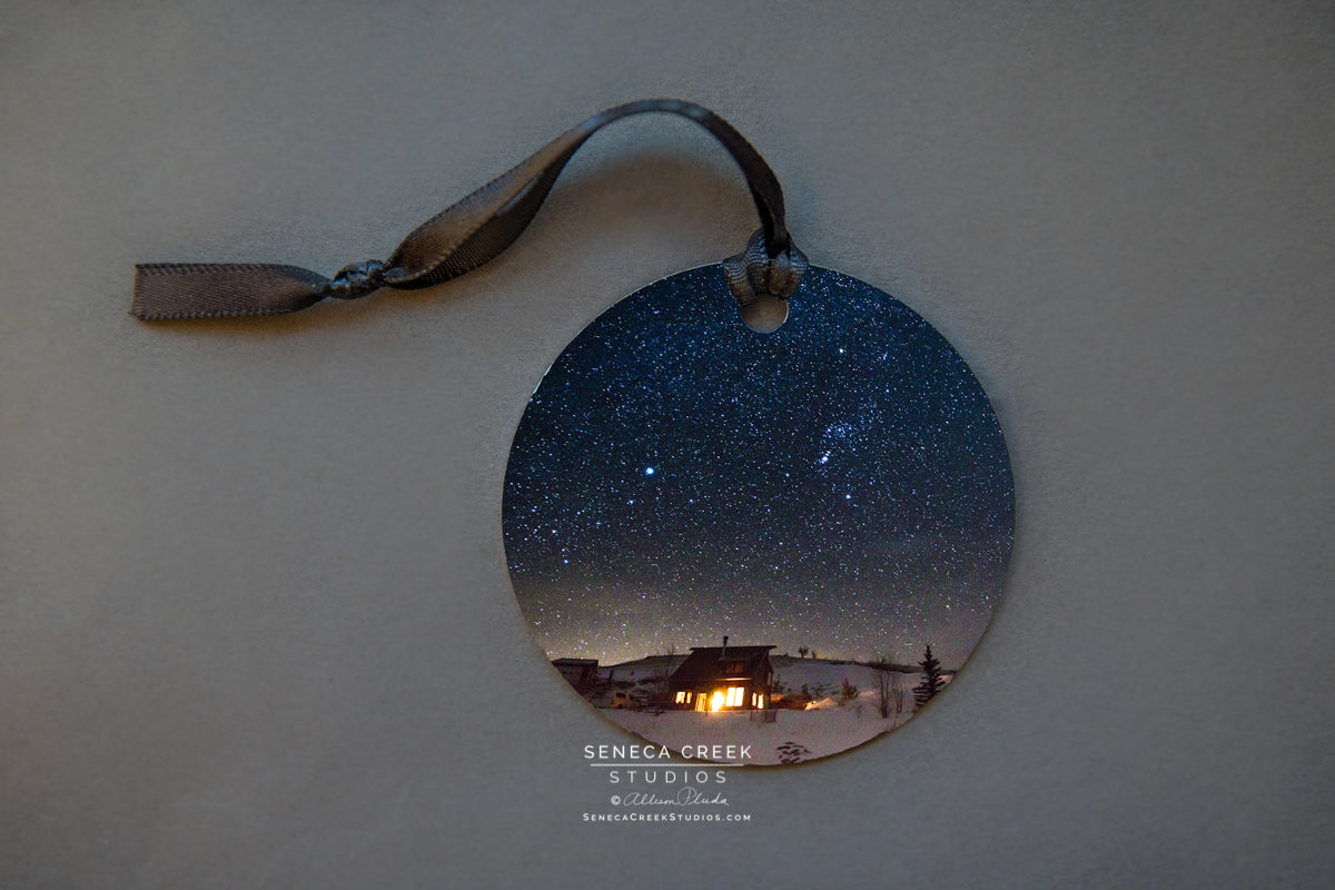 Starry Cabin Metal Print Ornament Photographer: Allison Pluda, Seneca Creek Studios A hanging photograph metal ornament A photographic picture of a starry cabin up in the Snowy Range mountains Picture on both sides of the ornament and the ornament hangs with a ribbon Please note, each photograph for the ornament has been taken by the Artist
