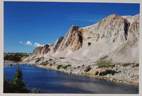 Lookout Lake " 10,627 Feet in Elevation " the Snowy Range Artist: Susan Davis  Printed with pigment ink on metallic paper  Looking from Left to Right:  The Diamond at 11,720 feet; Pillar Buttress, Sundial Slab at 11,740 feet; Old Main and Triangle Buttress at 11,755 feet  Lookout lake sits below the large face of the granite with pine trees along the bank  19" long x 13" high print  Foam board backing and in a plastic sleeve for added protection  Ready for a frame of your choice