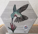 "Hummingbird" Hexagonal Handpainted Trivets Artist: Beth Rulli  This Trivet is a repurposed ceramic tile  Cork on back of tile protects table surface from scratching  Decorative or Functional  Hand painted color  Screen printed  Hummingbird feeding off an Indian Paintbrush flower, which is Wyoming's State Flower.  Approximately Size 8 1/4" x 7 1/4" x 1/2"  Slight variations in shade of color  This piece is custom designed by the Artist , with a slight variation between each piece
