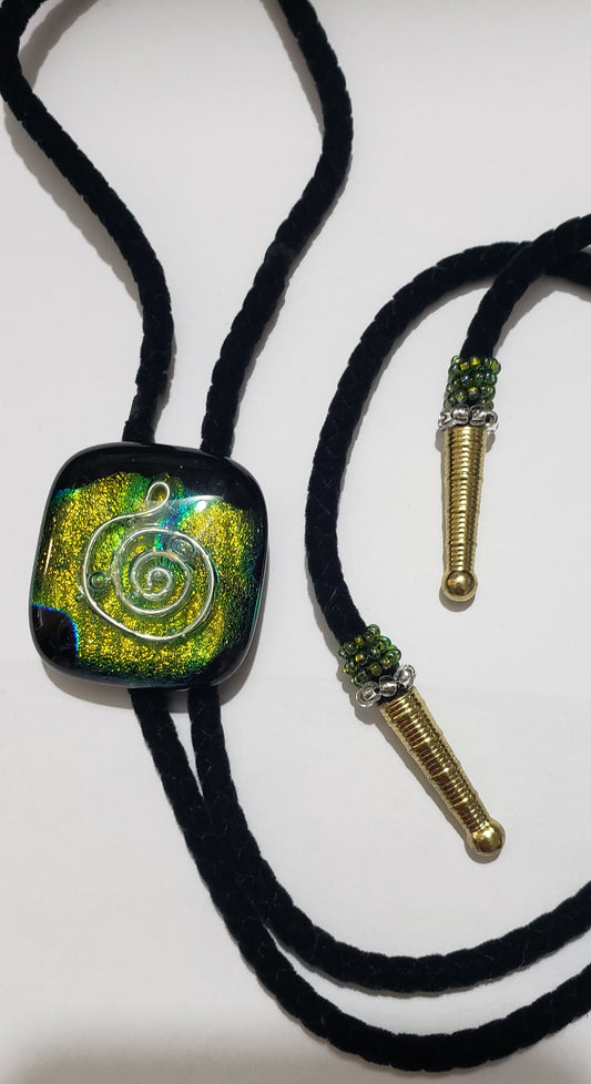  Bolo Tie with Custom Dichroic Fused Glass Pendant Fused Glass Artist: Leslie Irving  1 3/8" x 1 3/8 "  Custom made glass pendant with iridescent blues / greens   Soft black cloth bolo tie 16" length  Gold metal tips  Custom beading at the bolo tips