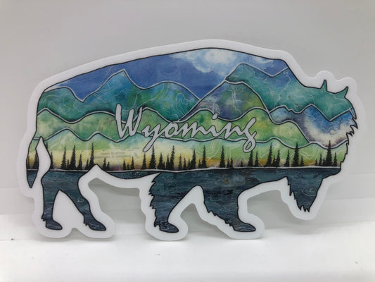 " Wyoming Buffalo " Sticker Artist: Nancy Marlatt  Shaped Cutout Vinyl Sticker  4.75 " length  x  3" wide  Mixed media design of a lake, forest line, mountains and skies of Wyoming  Wyoming is written across the sticker  Print from an original work by the artist  Great to use on a water bottle, laptop, or anywhere for decoration  Made from Nancy Marlatt's original artwork  