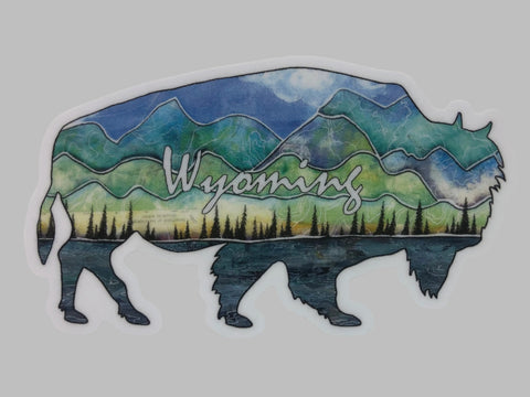 " Wyoming Buffalo " Sticker Artist: Nancy Marlatt  Shaped Cutout Vinyl Sticker  4.75 " length  x  3" wide  Mixed media design of a lake, forest line, mountains and skies of Wyoming  Wyoming is written across the sticker  Print from an original work by the artist  Great to use on a water bottle, laptop, or anywhere for decoration  Made from Nancy Marlatt's original artwork  