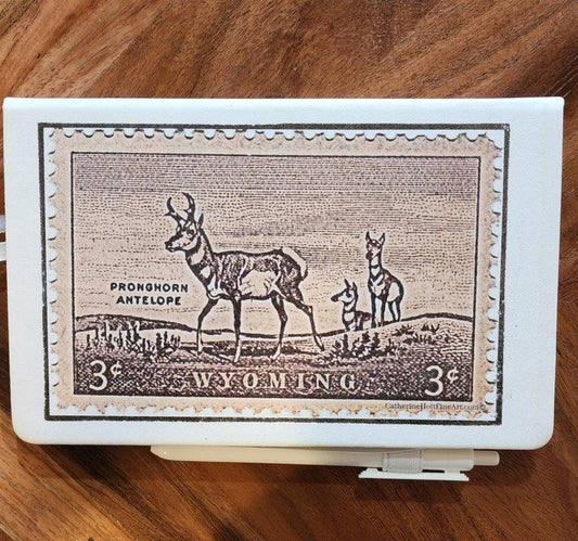 " 3 Cent Pronghorn Stamp " White Journal with Pen Artist: Catherine Holt  White leather covered journal  3 Cent Pronghorn Antelope Stamp cover art   Cover art is from an original painting by the artist  Lined pages with ribbon to mark your page  Pen and elastic pen loop included  Elastic band helps hold journal secure  4.5" long x 5.5" high  &nbsp;A beautiful journal for everyday writing and note keeping