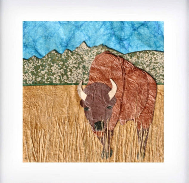 matted print of a paper collage art of a bison standing in grass with mountains a blue sky behind
