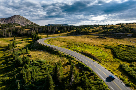 A drone aerial view of the Snowy Range Scenic Highway, highway 130, as it winds below Sugarloaf Mountain in the Medicine Bow National Forest  A blue car travels along the highway, giving a glimpse of the vastness of the Snowy Range