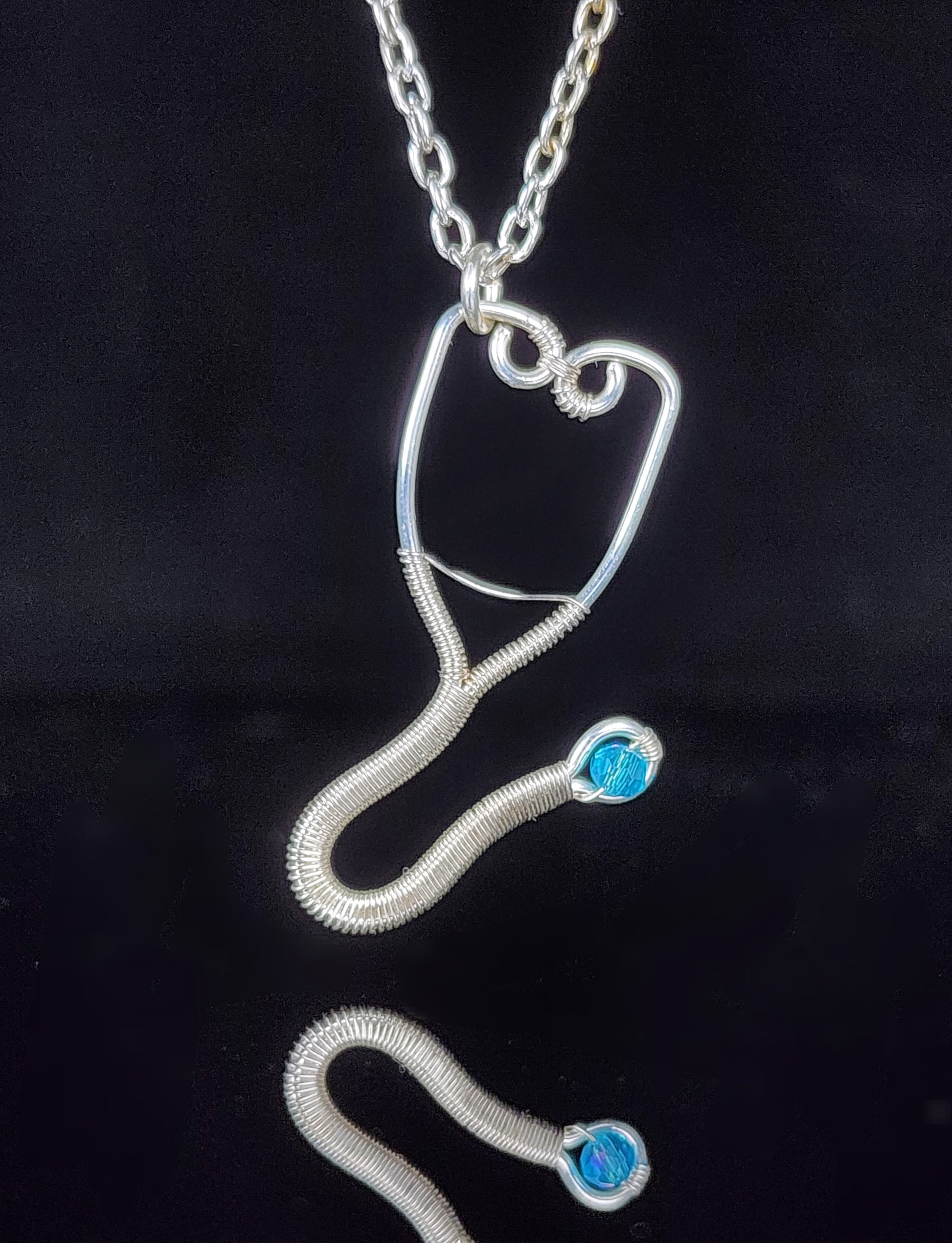 Hand woven silver colored artistic wire  Stethoscope design  Blue Swarovski bead as the diaphragm portion of the stethoscope  1.44" long x .8" wide  Silver colored chain necklace with lobster clasp  Care instructions and cleaning cloth provided