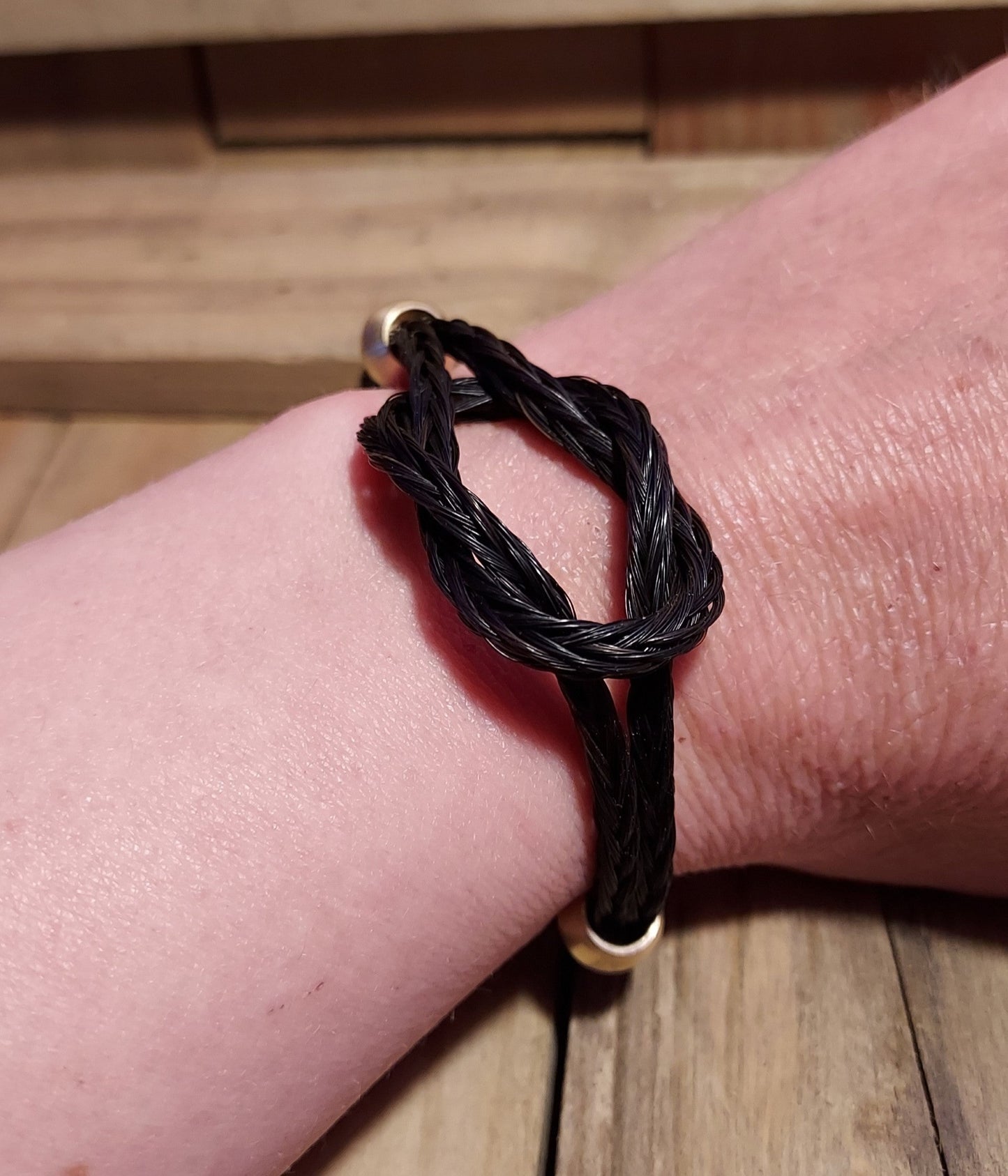 hand braided black horsehair linked to resemble an infinity knot and has two brass beads along each side. Bracelet is shown on a wrist