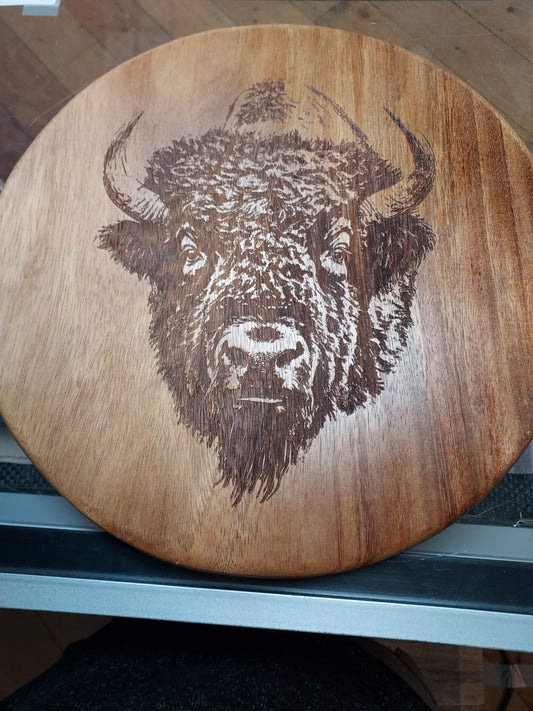 " Bison " Laser Engraved Round Charcuterie Board&nbsp;
Artist: Marcy Knotwell

Elevate your entertaining and gifting with this stunning East Indian Walnut Charcuterie Board!

Handcrafted and engraved with a majestic bison head, this versatile board is perfect for serving cheese, charcuterie, snacks, or using as a trivet. It's also functional for cutting herbs, lemons, limes, and garlic

Features: Solid East Indian Walnut: Beautiful, durable, and naturally antimicrobial
Handcrafted &amp; Engraved: 