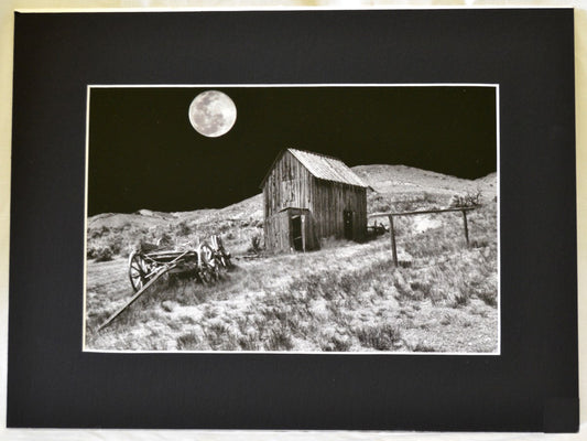 Matted Black and White photograph of an old barn, old wagon, and a full moon  The image was taken in Bannack State Park, Montana  2" wide black matting with white edging around the print  12" long x 8" high print  16" long x 12" high as matted  In a plastic sleeve and on sturdy foam board for added protection  Ready for a frame or to be displayed on an easel