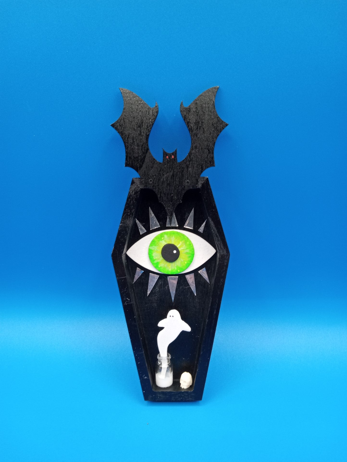 Hand painted wooden coffin figure with bat figure on the top  Large green eye peaking out of the coffin  Ghost coming out of a bottle and a small skull off to the side  Wire attached to the back for hanging