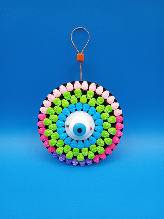 Colorful skull mandala  Pink, green, purple and blue skulls attached to black wooden board  Blue eye in the center of the mandala  Wire hanger on the back  This mandala is looking back at you  ( Eye See You )