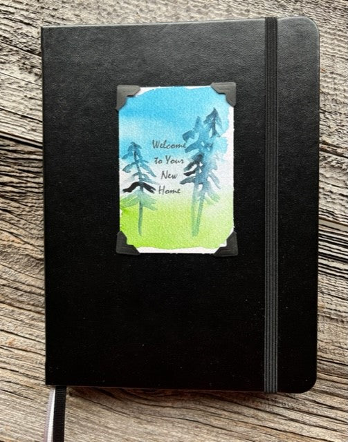 Hardcover black lined journal  Black elastic band helps hold the book closed  Ribbon page marker  Welcome to Your New Home watercolor with pine trees, blue sky and green grass  Lined pages inside  A beautiful journal for young and old alike  6" long x 8" high x 3/4" wide