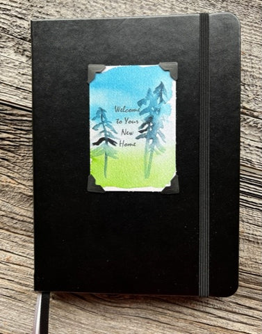 Hardcover black lined journal  Black elastic band helps hold the book closed  Ribbon page marker  Welcome to Your New Home watercolor with pine trees, blue sky and green grass  Lined pages inside  A beautiful journal for young and old alike  6" long x 8" high x 3/4" wide