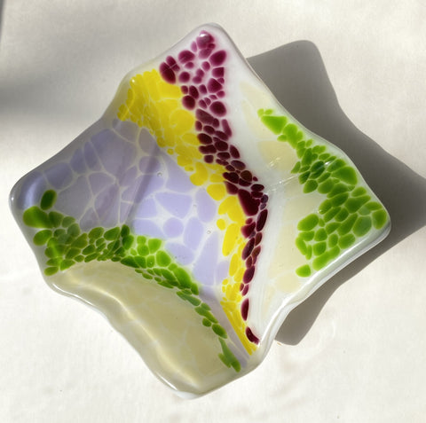 " Pathways " Fused Glass Candy Dish Artist: Shari Wolf Fused glass dish  Squarish shape with folded corner edges  Green, purple, yellow and lilac colored pieces of glass patterned into pathways on a cream colored glass background  5" long x 5" wide x 2.5" high  Use as a candy dish or to hold other small items like a candle, keys, or coins  Attach a plate holder and you can hang the dish from the wall as art, or in the window as a suncatcher  A beautiful multi-faceted dish