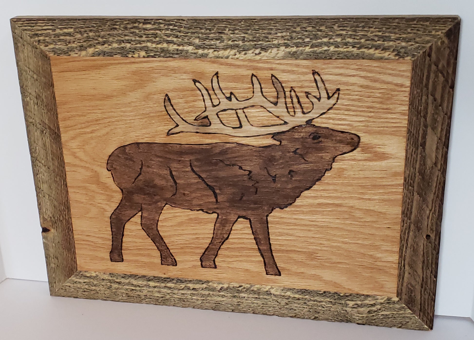Wood Burned Elk Framed Wall Art Artist: Michael McMahon  Hardwood plywood sanded and burned  Detailed Elk outline stained in walnut  Golden oak background  Polyurethane coated  Rough cut wooden frame stained in brown  Wire hanger on the back  19" long x 14.5" high x .75" wide  Would be perfect for a cabin or for any outdoorsman