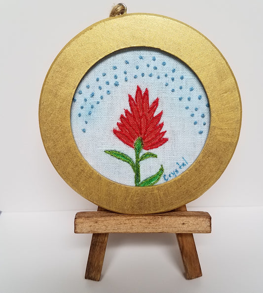Round Indian Paintbrush Fiber Art Ornament Artist: Crystal Lawrence Hand painted and hand embellished Indian Paintbrush on a blue background  4" across  Cute golden round frame for hanging  Easel is included  Would make a wonderful conversation starter   Please note, each piece is a handmade custom designed by the Artist , with a slight variation between each piece