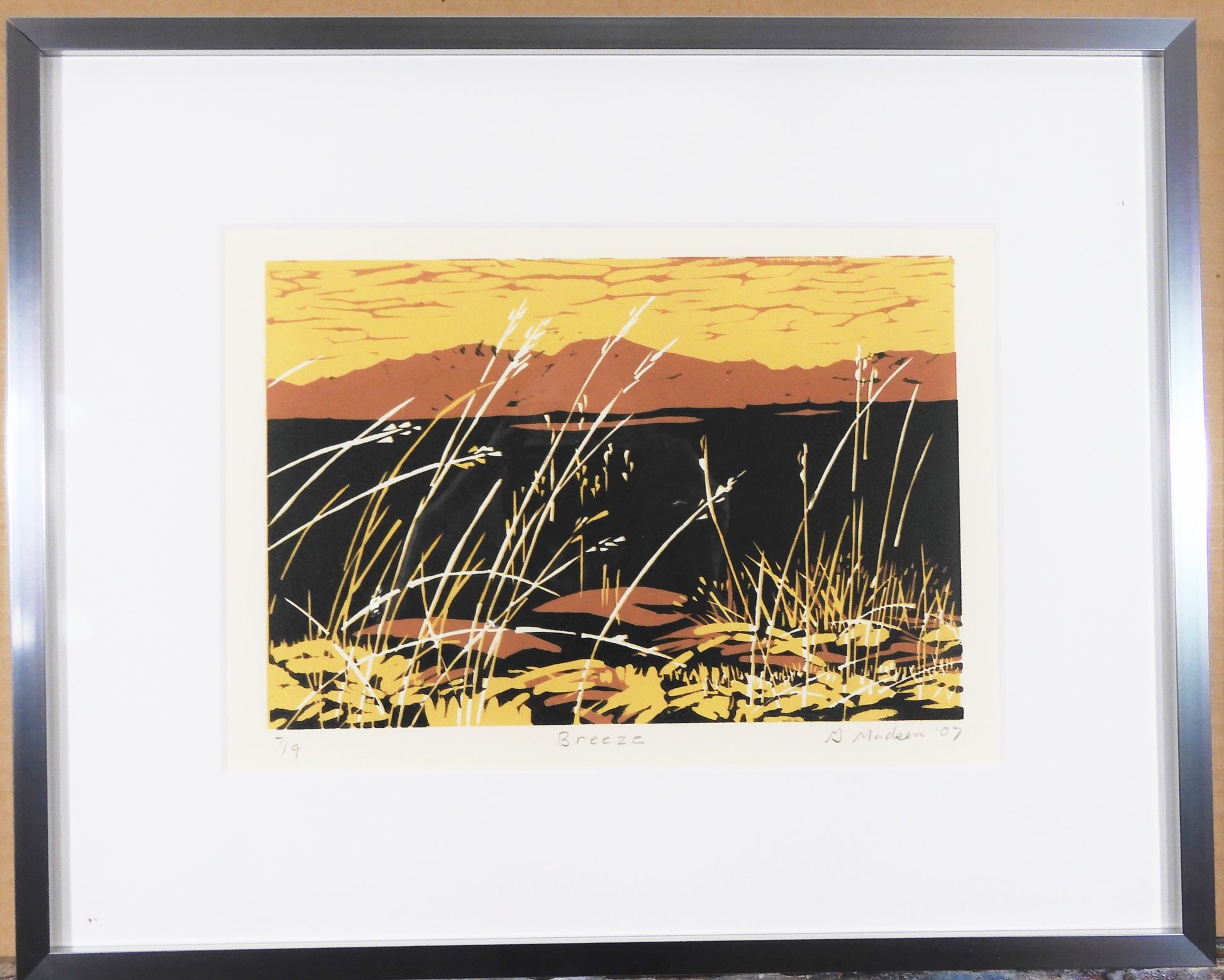 silver metal framed relief print. grasses waving in the breeze in the foreground. Orange mountain silhouette in the background.