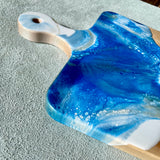 Rounded Edge Cheese Board / Handle Artist: Marcy Knotwell  Blue, White with liquid gold lacing resin design  You will not be disappointed with the quality of this product! Our handcrafted wood grained-edge charcuterie cheese boards are the answer to stylish, multi-functional, sustainable serve-ware.  When not in use, this charcuterie cheese board makes a beautiful addition to your countertop or gallery wall.