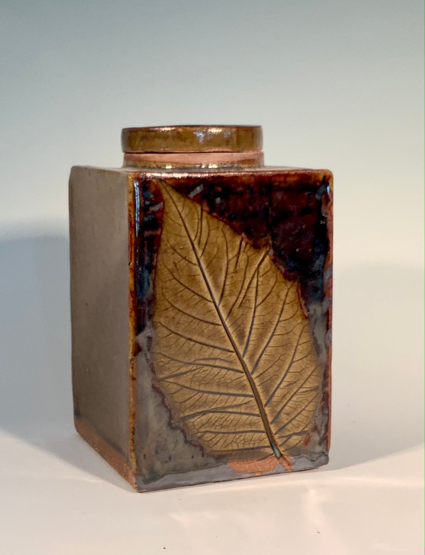 Rectangle Stoneware Jar / Leaf Imprint Muffy Moore: Ceramic Potter Stoneware jar with green / brown glaze  Leaf imprint on two sides with round stoneware lid     3 1/2" Long x 3 1/2" Wide x 6" Tall  Beautiful hand thrown stoneware jar with lid   Dishwasher, oven and food safe  Great for storing loose leaf teas or coffee