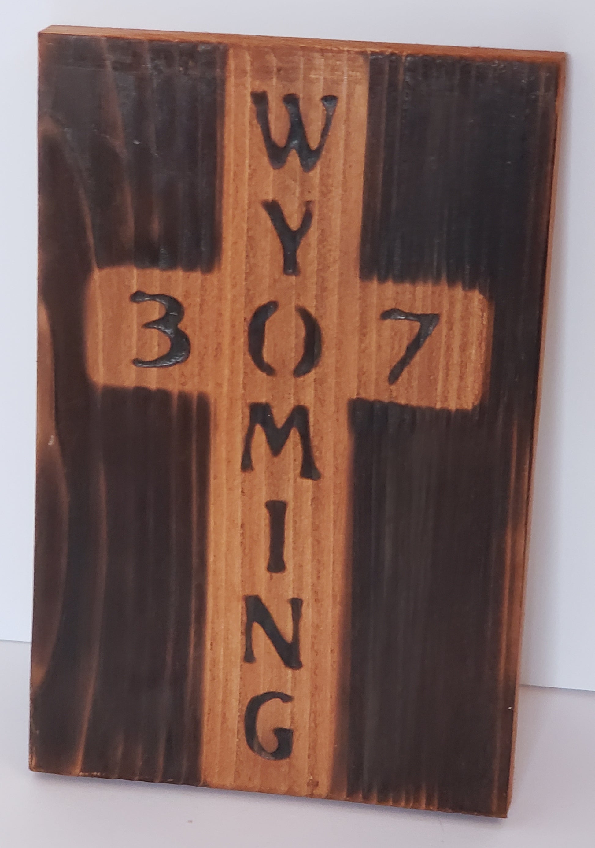 Wyoming and 307 Wood Burned Cross Artist: Michael McMahon  8 1/4" long x 5 1/4" wide x 1/2" high  Rectangle cedar board with the word Wyoming and 307 burned into the board  Wood frame  burned and wire brushed color and clear protective polyurethane finish  Wire hanger on the back  Wood stain color may vary slightly.