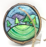 " Hill Side " Hand Painted Wooden Ornament Artist: Nancy Marlatt  Hand painted natural wood slice with a jute string attached  Original watercolor painting  Actual size and weight varies per piece  Hand signed by the artist  Green grass in the foreground  A row of pine trees  Hills in the background  Clouds in the sky  2.5" x2.53" .25"
