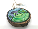 " Hill Side " Hand Painted Wooden Ornament Artist: Nancy Marlatt  Hand painted natural wood slice with a jute string attached. Green grass in the foreground  A row of pine trees  Hills in the background  Clouds in the sky. Original painting by Nancy Marlatt, Wyoming