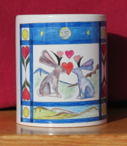 8 oz white ceramic mug with handle  Design is from an original drawing by the artist  Two Jackalope with hearts between them and mountains in the background  Blue boarder with hearts, greenery, along with moon, mountains, and stars  Add a little packet of chocolate kisses and make a perfect Valentine's gift to the one you love