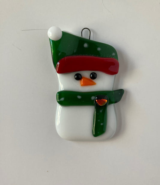 " Christmas Spirit " Snowman Ornament Artist: Shari Wolf Fused Glass Ornament  Snowman in a dark green hat and scarf with white dots  Red trim and white pompom on the hat with a jewel on the scarf  2" long x 3" high x 1/4" wide fused glass  Wire hook attached for hanging  Great for a Christmas Tree or just as a fun Holiday Decoration